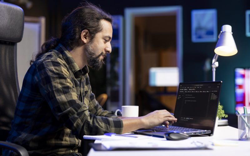 Male programmer works on his portable computer, managing the database and monitoring network for cyber security. Young man sitting at desk and using digital laptop for coding.